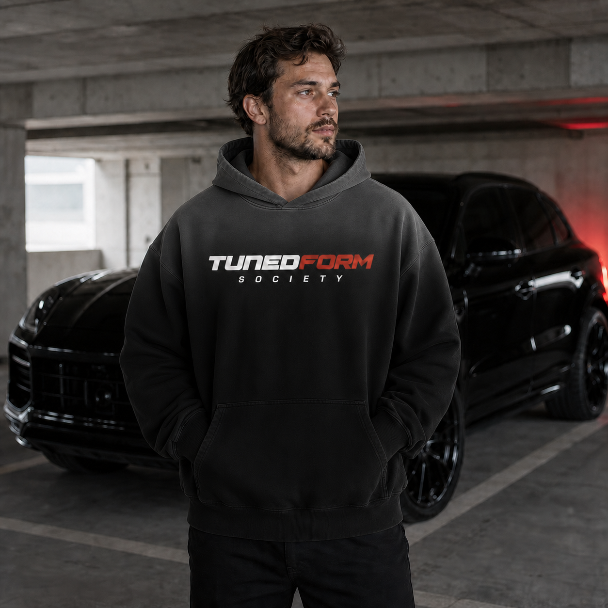 Male model wearing the black TunedForm Society heavyweight hoodie with a charcoal sunfade gradient across the hood and upper chest beside a black performance SUV